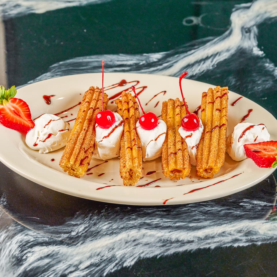 Plate of cinnamon-sugar churros with whipped cream, cherries, strawberries, and chocolate drizzle.