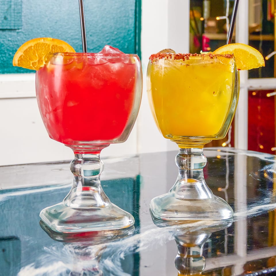 Two colorful iced drinks in stemmed glasses, garnished with orange slices and straws, sit on a reflective restaurant table.