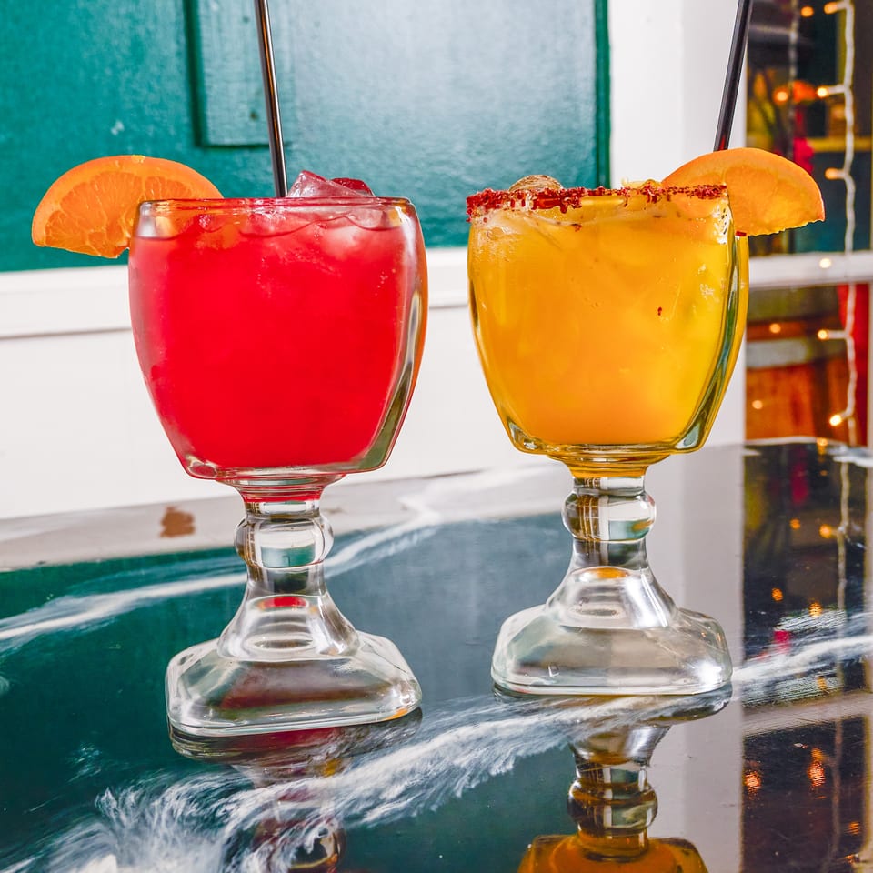 Two colorful cocktails with ice, orange garnishes, and chili-salt rims sit in stemmed glasses on a reflective restaurant table.