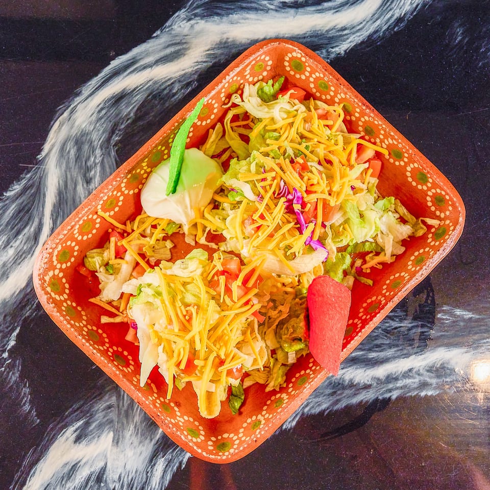 Colorful taco salad with lettuce, tomatoes, shredded cheese, guacamole, sour cream, and tortilla chips in a decorative red bowl.