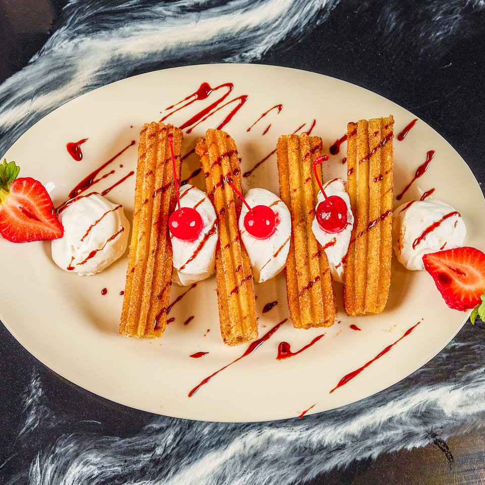 Golden churros with whipped cream, cherries, strawberries, and chocolate drizzle arranged on a large oval plate.